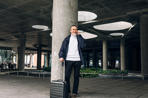 Smiling mature traveller standing outdoors with luggage and talking on cell phone