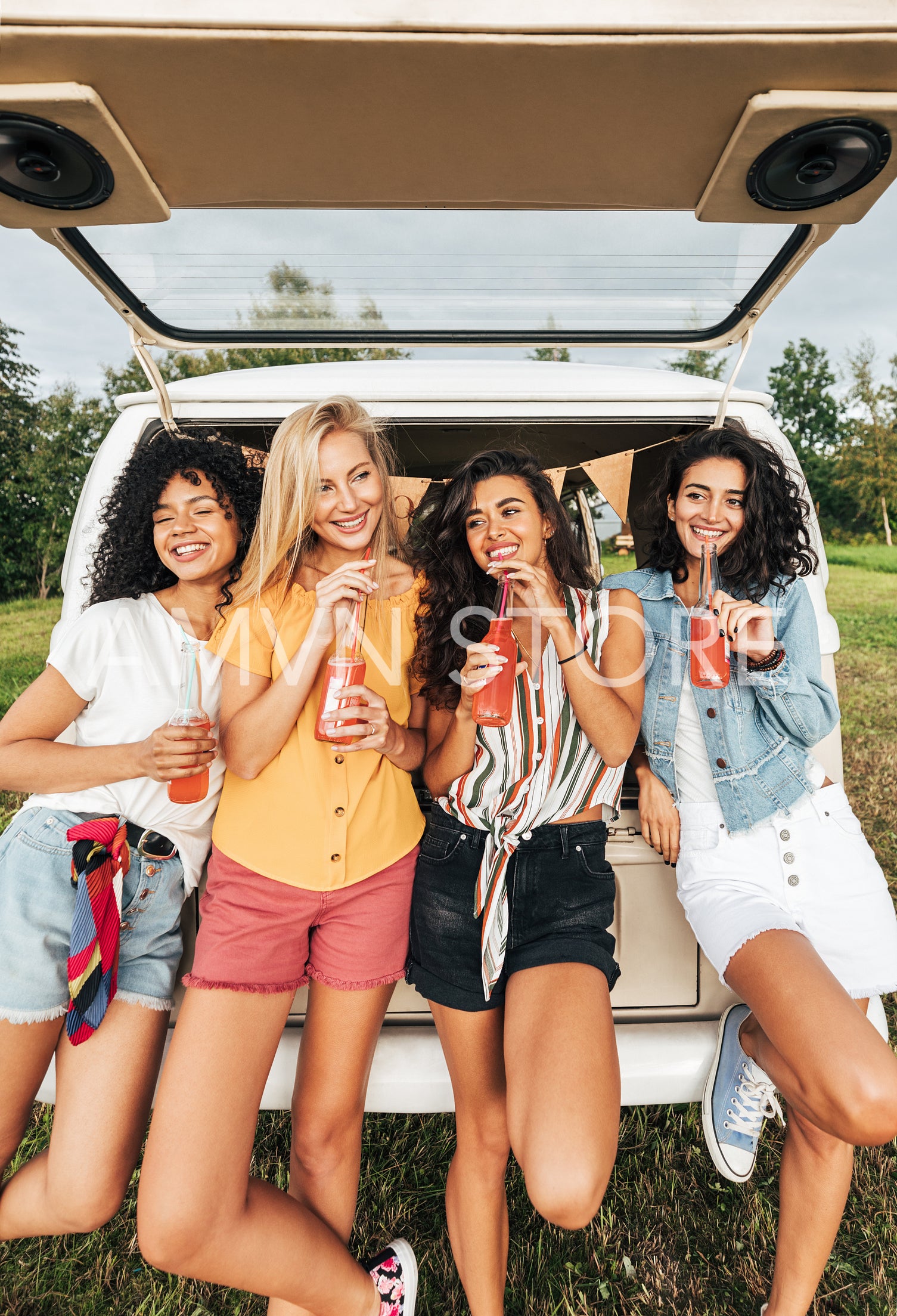 Group of diverse women holding cocktails while standing at the back of camper van. Four females with bottles standing at a car during summer vacation.