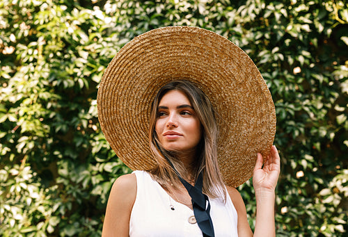 Young woman wearing a straw hat standing outdoors in front of a wall with green leaves