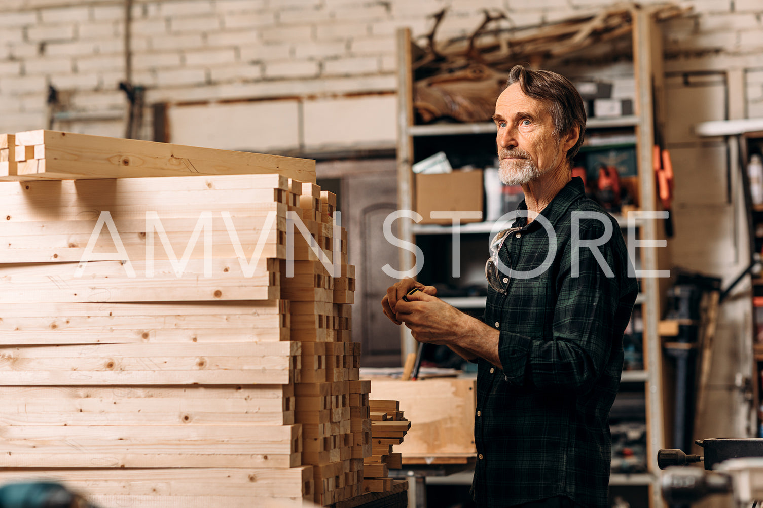 Senior man standing near a bunch wooden parts of furniture in a workshop and looking away	