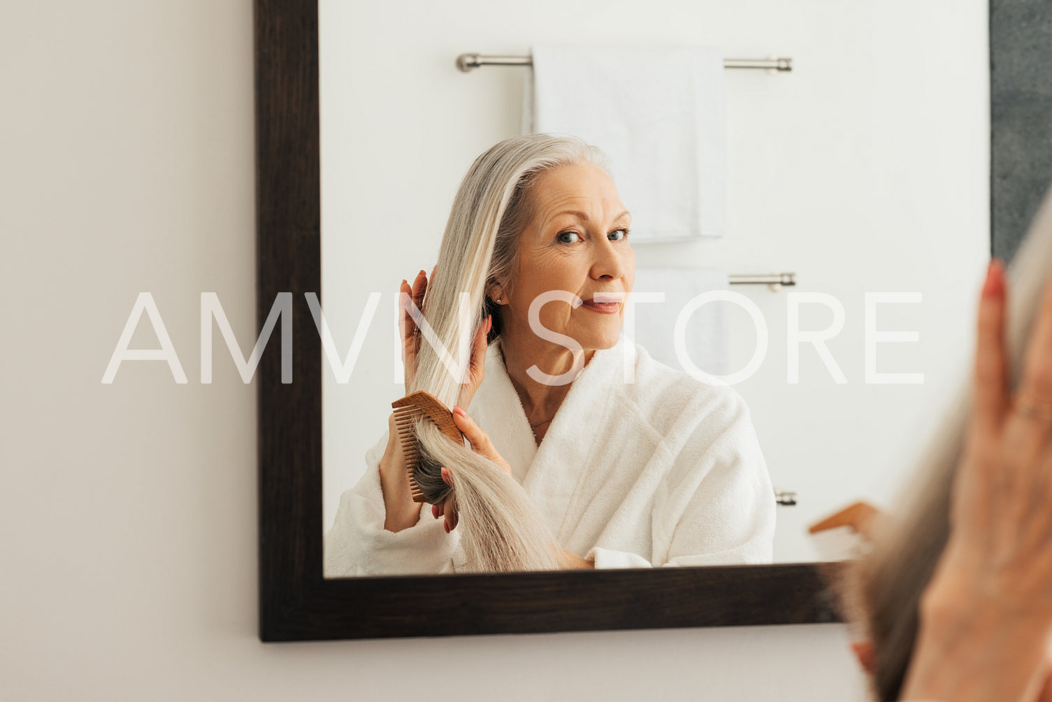 Senior woman looking at mirror while combing her long hair in the bathroom