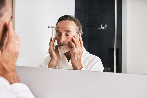 Senior man massaging his face while standing in front of a mirror in bathroom