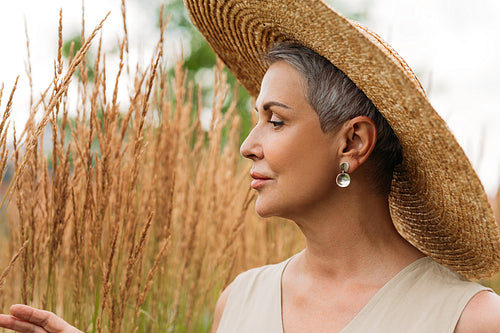 Side view of a senior woman in straw hat looking at wheat on a field