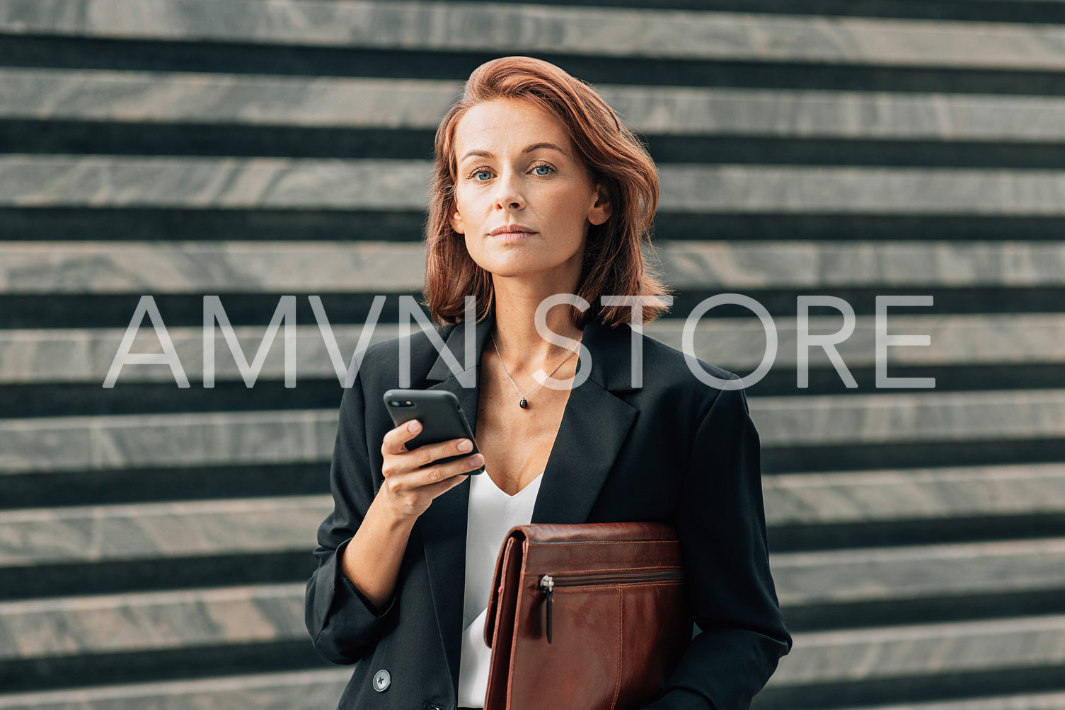 Middle-aged businesswoman holding a leather folder and smartphone and looking at camera while standing outdoors