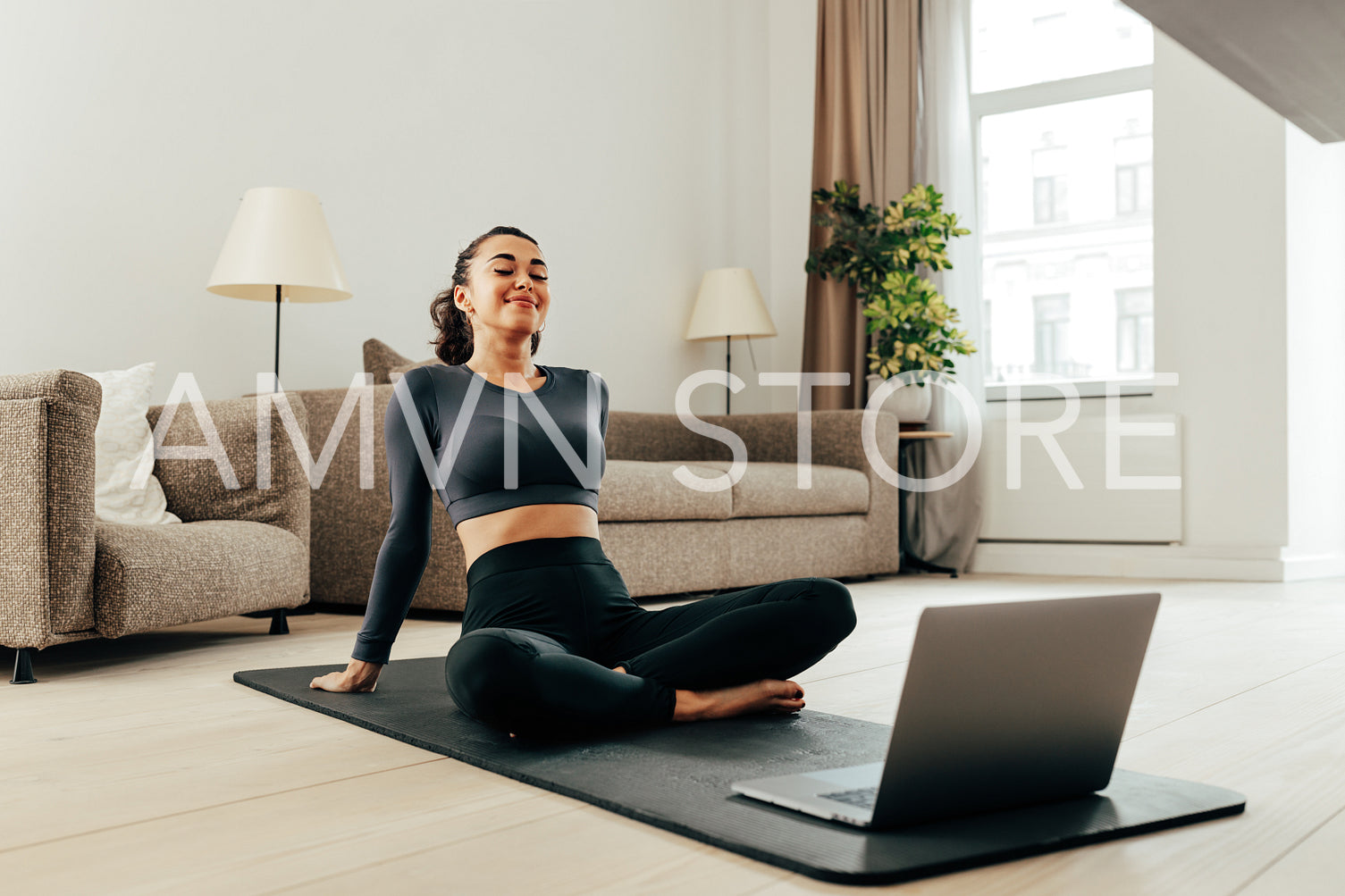 Smiling woman in sport clothes sitting on a mat in living room in front of a laptop. Young female relaxing during training at home.	