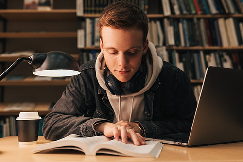 Student in casual clothes preparing exams late evening in library. Young male reading from book at desk.