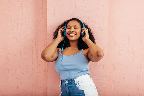 Woman wearing blue headphones enjoying music while standing with close eyes at pink wall