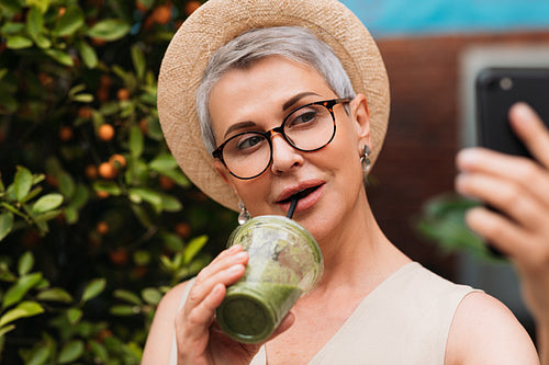 Senior woman taking a selfie outdoors while drinking a green smoothie. Portrait of a mature female in a small straw hat taking a selfie in the park.