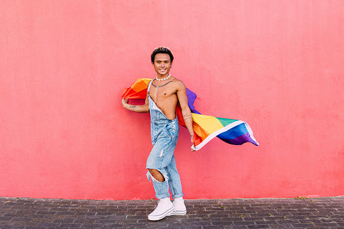 Smiling gay male walking with rainbow LGBT flag in city and looking at camera