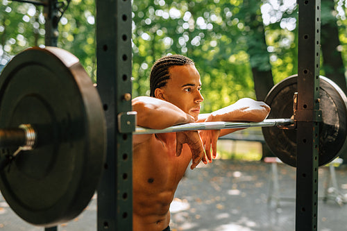 Side view of a bare-chest athlete standing outdoors leaning on a weight bar