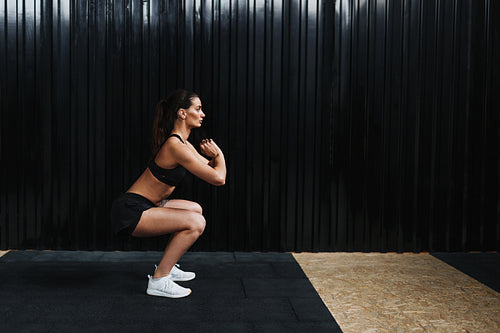 Side view of sporty woman practicing squat exercises in gym near black wall