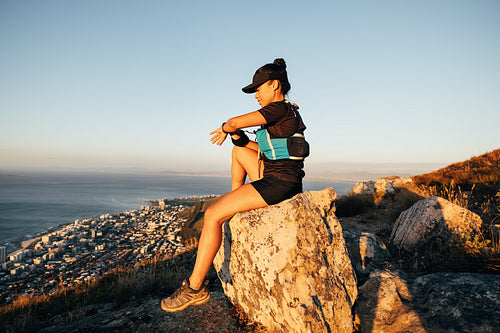 Side view of sports woman sitting on a rock checking smart watch at sunset. Trail runner relaxing on the top of the mountain.