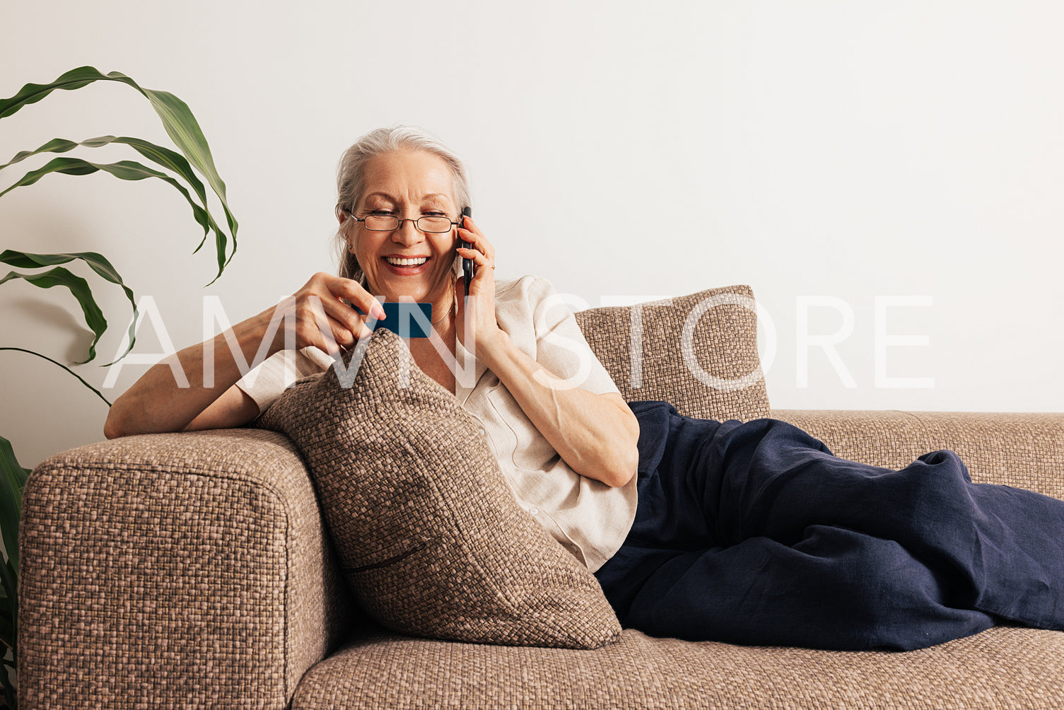 Happy senior woman holding a credit card and making call. Aged woman in glasses lying on sofa and shopping online.