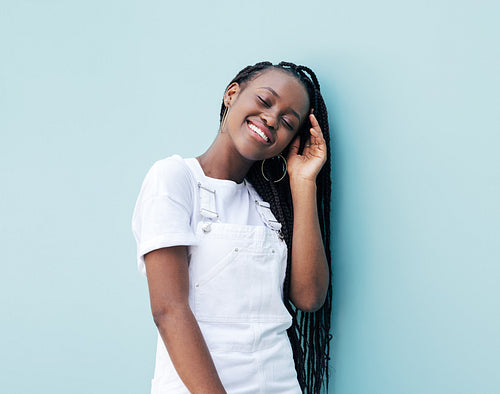 Cheerful female with braids wearing white clothes leaning on a blue wall with closed eyes