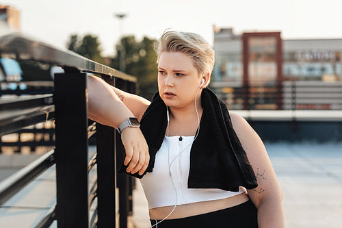 Curvy woman leaning on a railing on the roof. Young tired woman with towel on neck looking away.