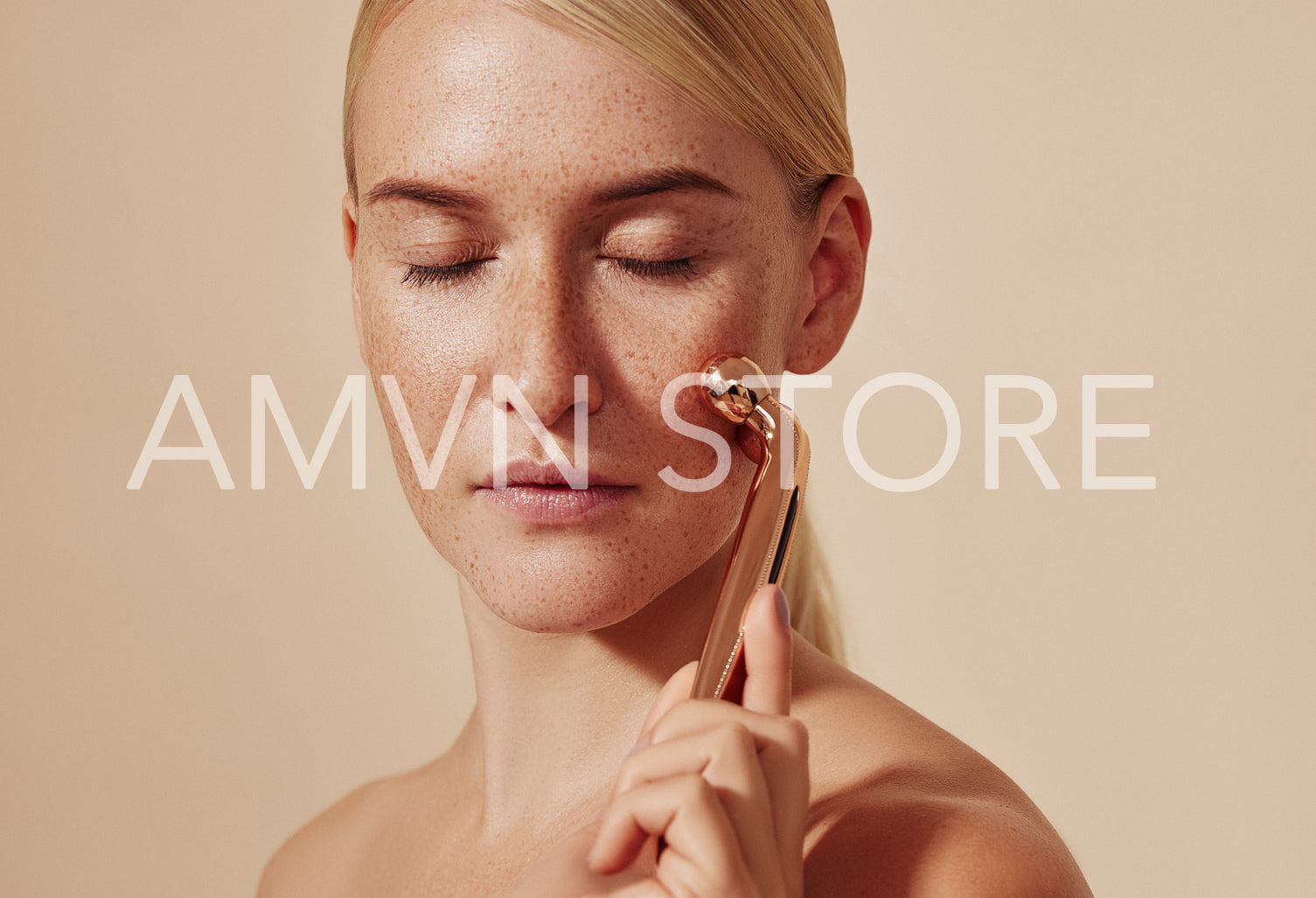 Young woman with closed eyes using face massager. Female with smooth freckled skin using face roller against a beige background.