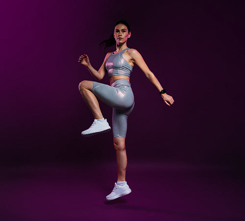 Full length of a slim female in silver fitness attire jumping up against a magenta background