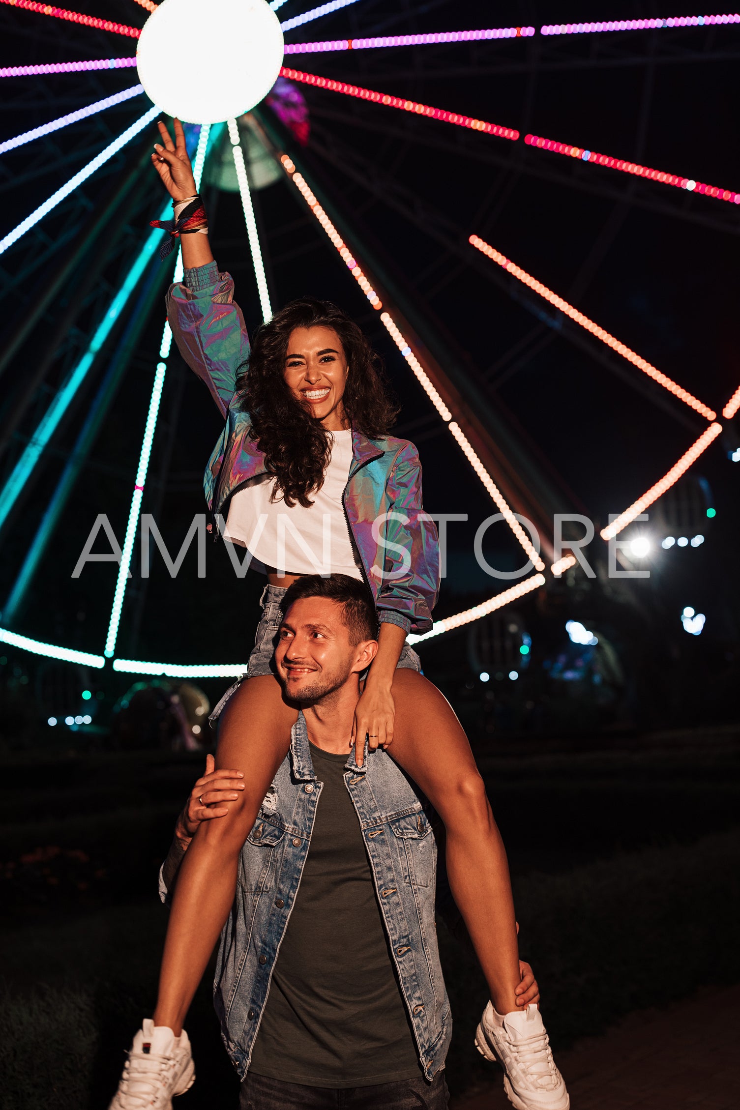 Smiling female sitting on the shoulders of her boyfriend and raising her hand in an amusement park at night