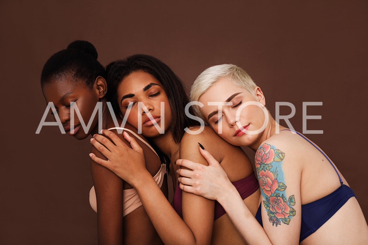 Three women in lingerie with closed eyes against a brown backdrop. Diverse females are embracing together.