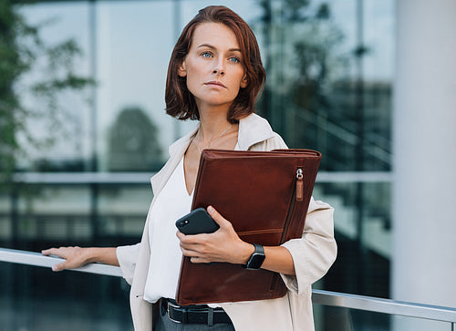 Confident middle-aged businesswoman looking away. Female in formal clothes holding leather folder while standing outdoors.