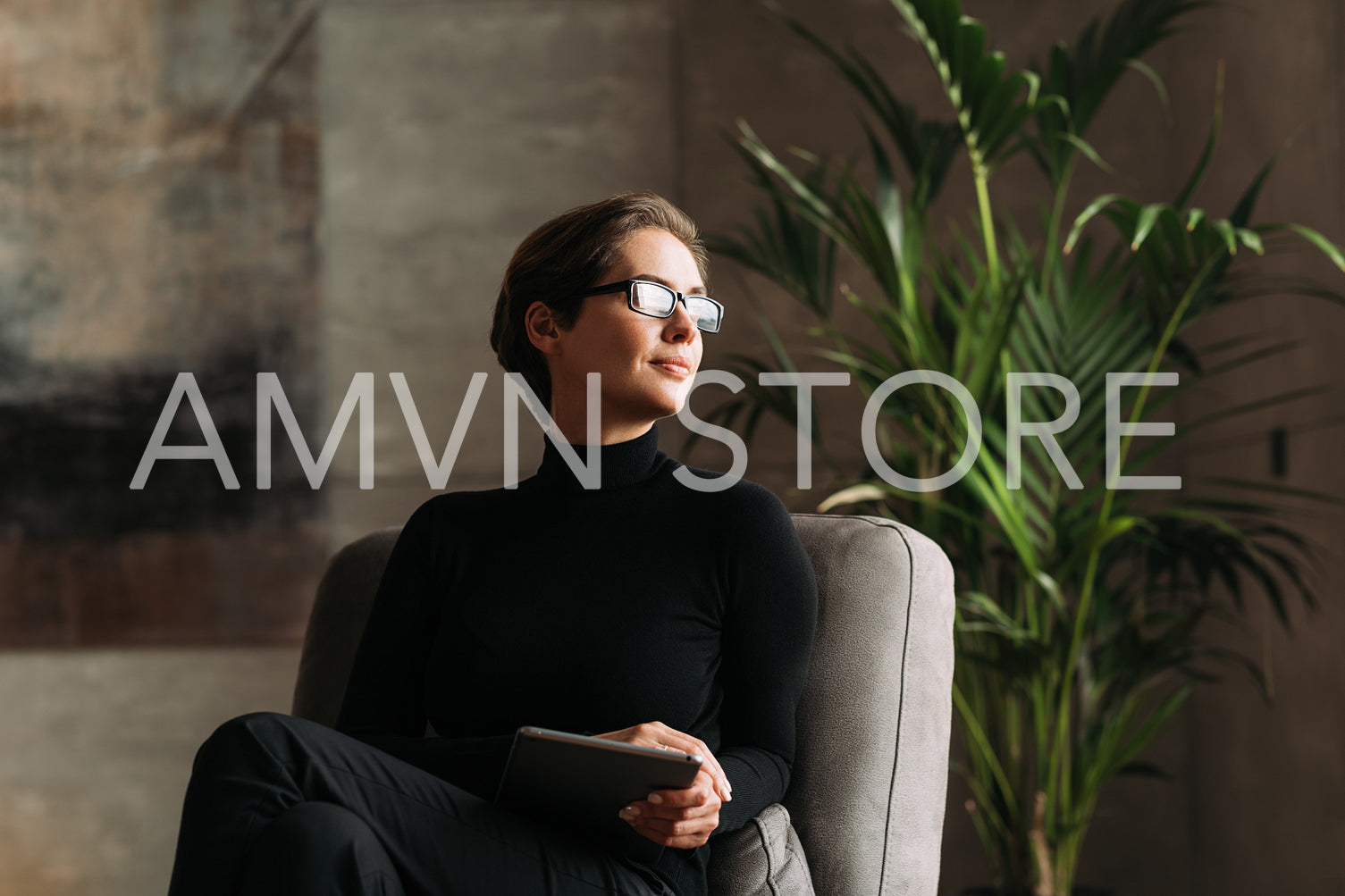 Young woman in black formal wear wearing glasses holding a digital tablet while sitting in her loft