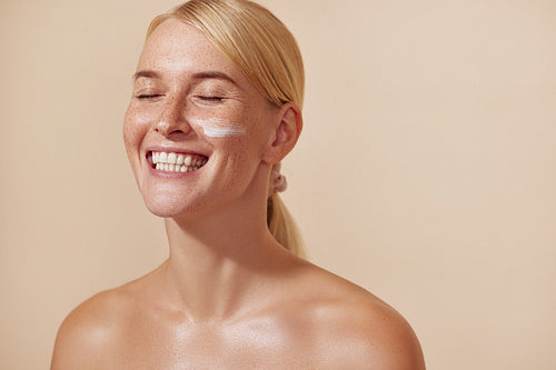 Happy woman with blonde hair and freckles. Young female with moisturizer on her cheek smiling with closed eyes in the studio.