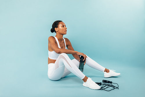 Tired woman in white sportswear sitting on a blue background. Female athlete holding water bottle in studio.