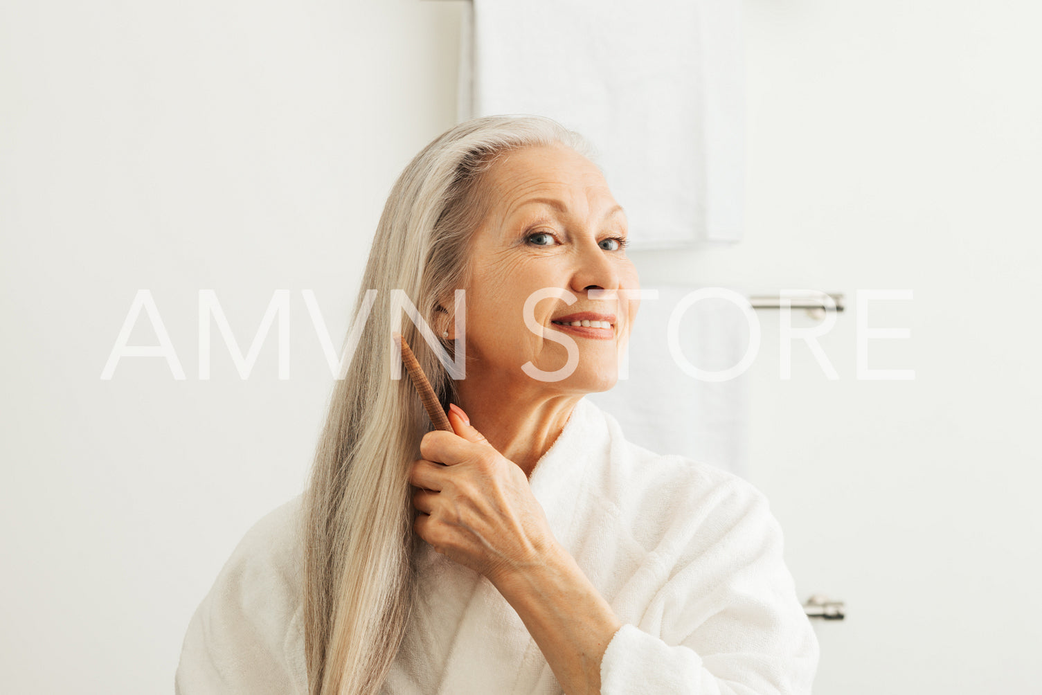 Senior adult woman combing her hair with a wood comb. Smiling female in a bathrobe taking care of her long gray hair.