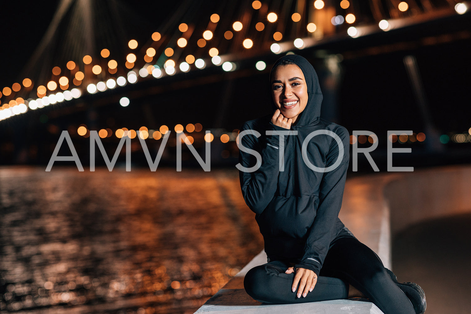 Beautiful middle east woman in hoodie sitting on embankment at night and resting after workout