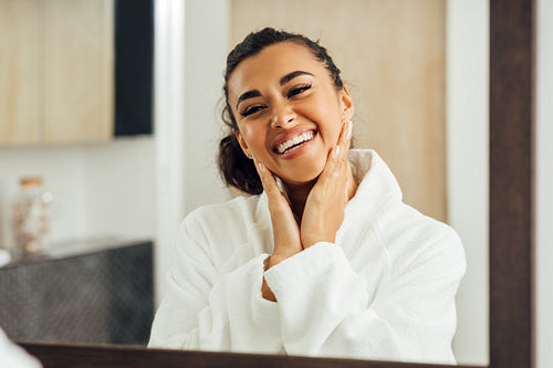 Happy woman in white bathrobe touching her cheeks. Young female looking in her reflection in bathroom mirror.