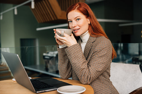 Young woman with ginger hair holding a cup. Female entrepreneur drinks coffee while working from a coffee shop.