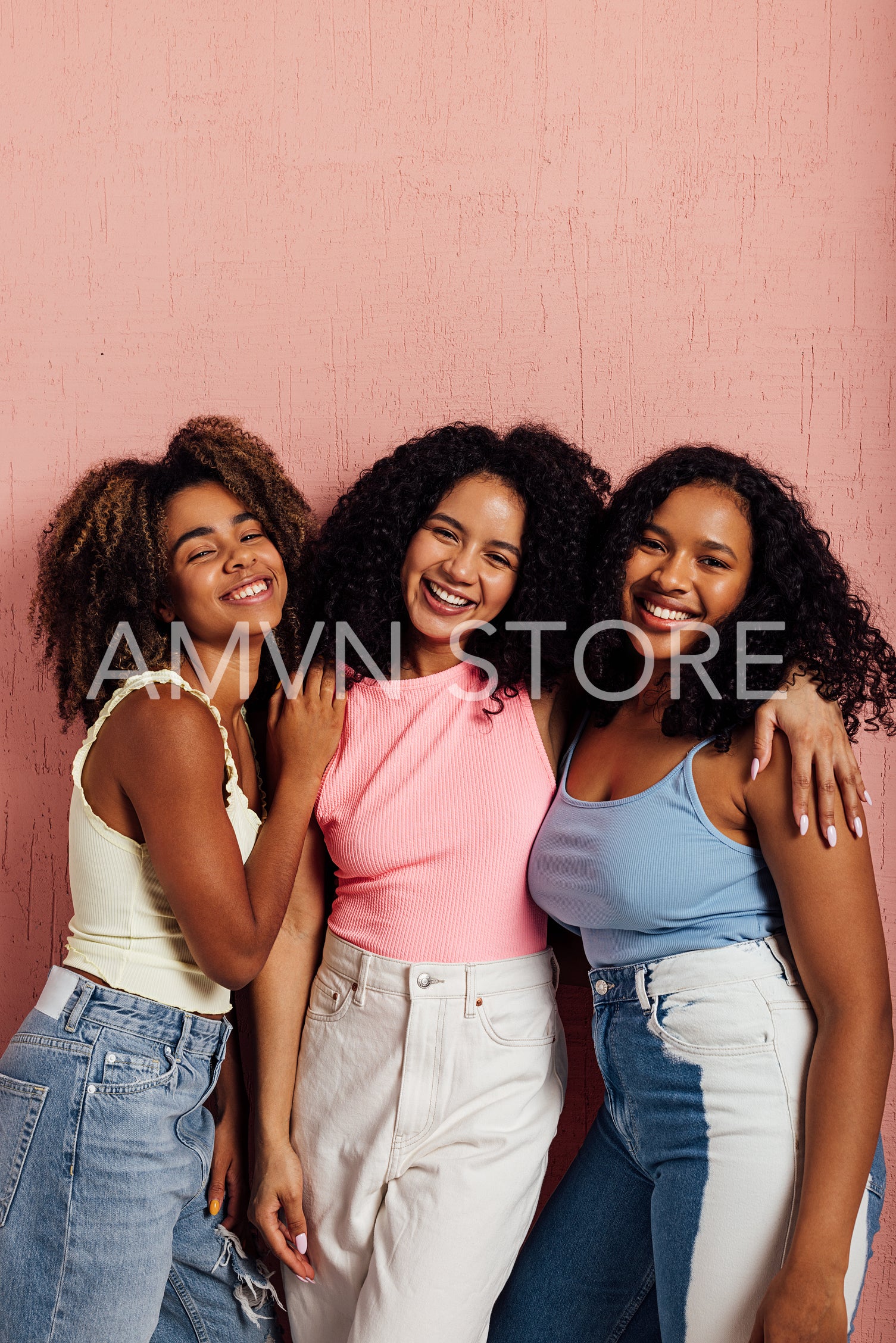 Three smiling women with curly hair standing together and looking at camera against a pink wall