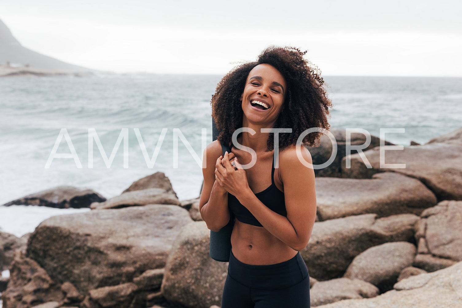 Laughing woman in fitness wear standing with closed eyes by seashore at sunset after training