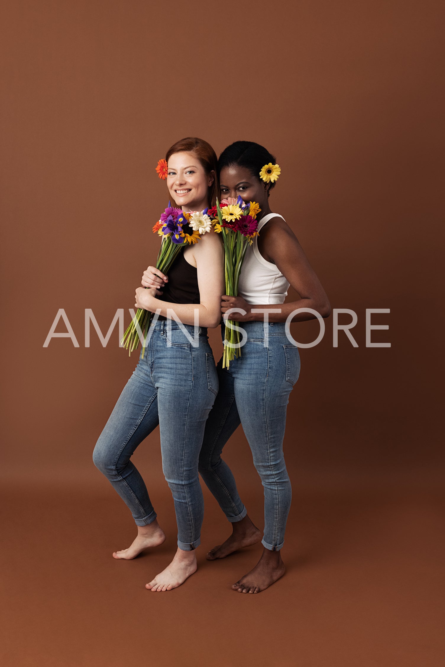 Two women with different skin color posing against a brown background. Cheerful females with flowers standing together looking at camera.