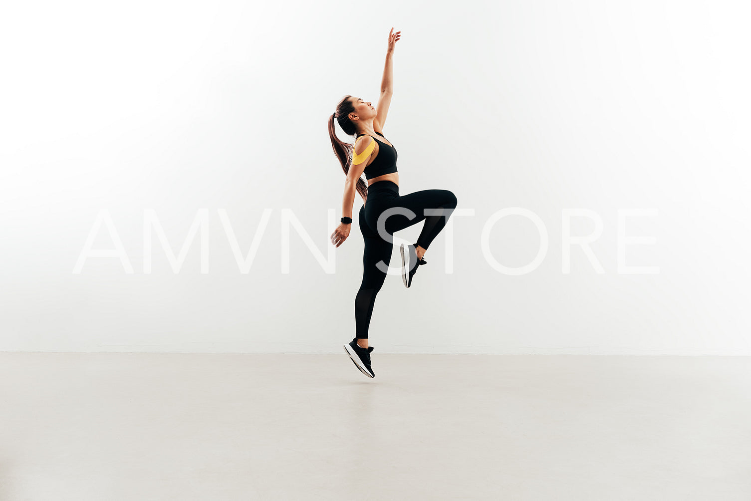 Woman jumping in the air against white wall raising one hand up. Young female in sports clothes warming up.