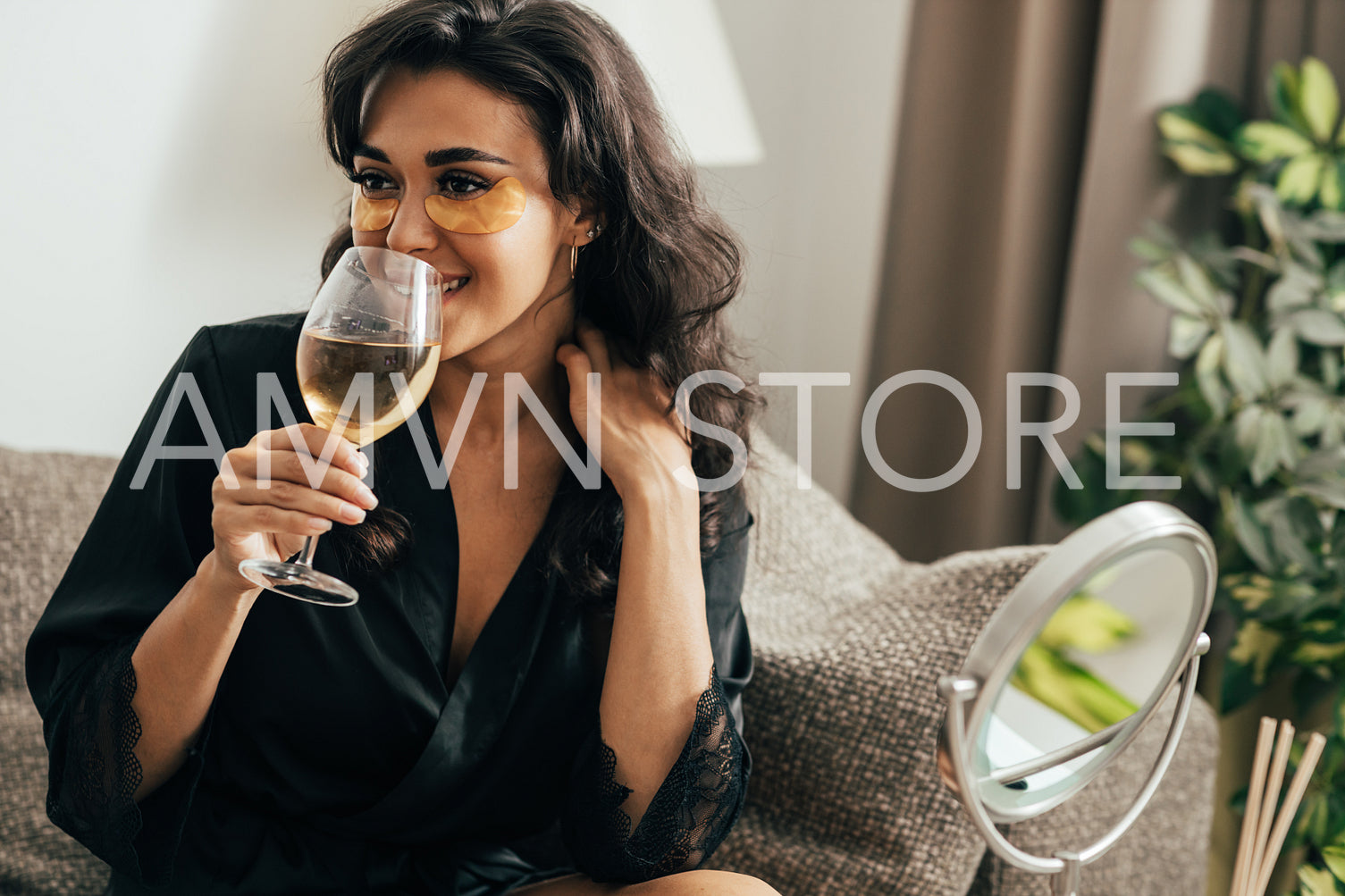 Brunette woman with eye patches drinking wine. Young female in satin robe relaxing at home on couch.	