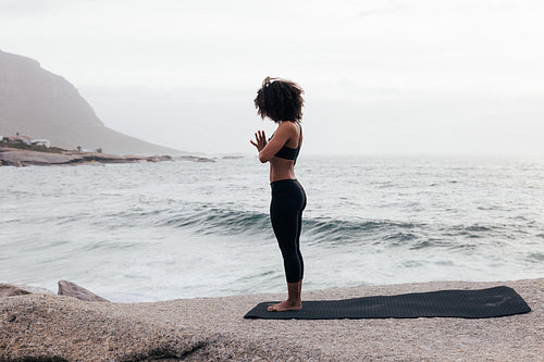 Side view of a slim woman meditating while standing on a mat against an ocean at evening