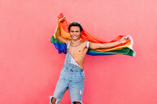 Happy male with LGBT flag standing against pink wall looking at camera