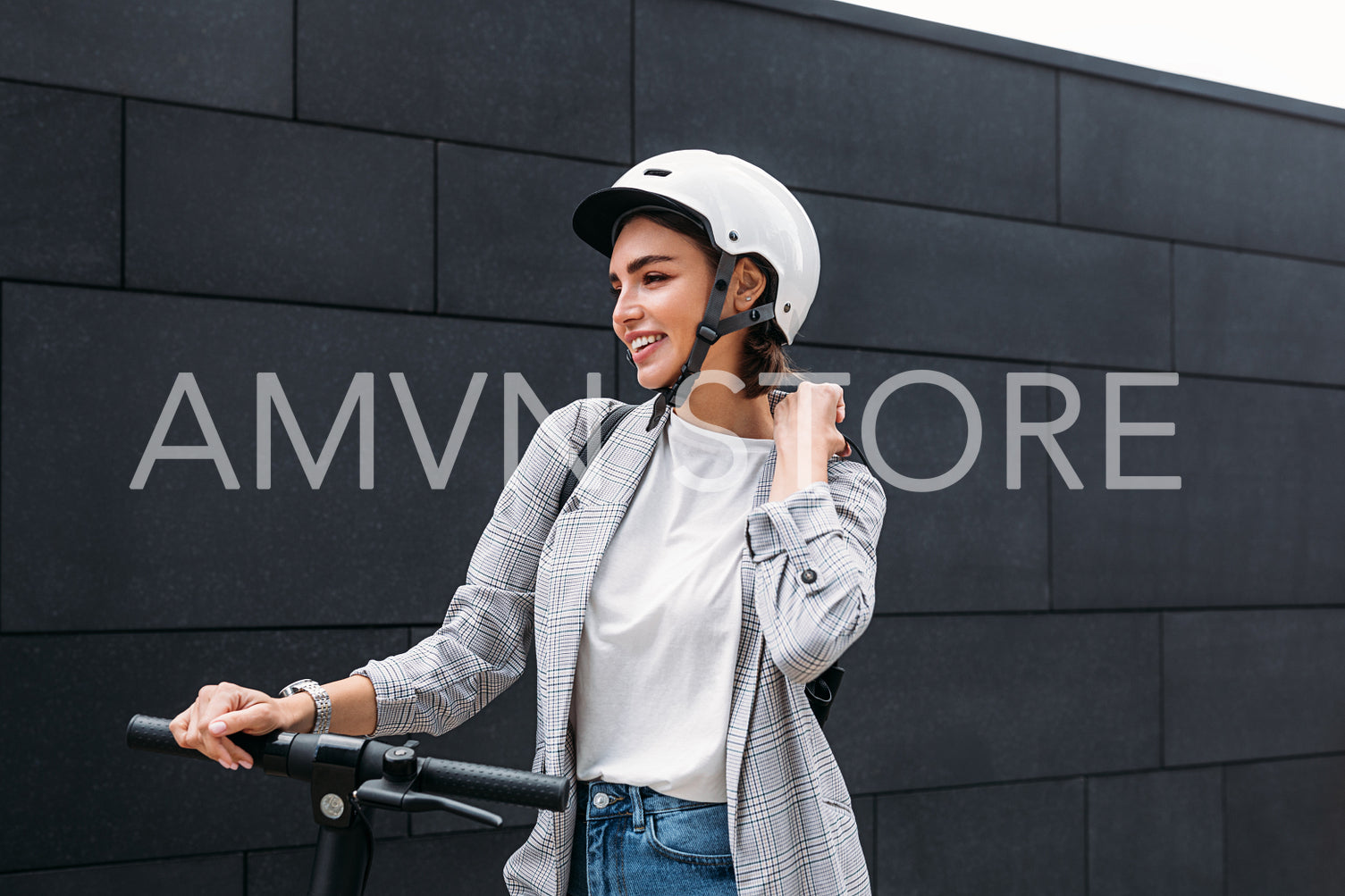 Young happy female adjusting strap of a backpack while standing at wall on electric scooter. Businesswoman with cycling helmet holding handlebar looking away.