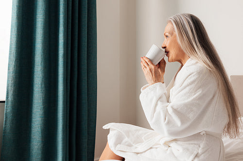 Senior woman drinking coffee in morning in bedroom. Female in bathrobe sitting on a bed looking at window.