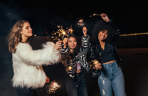 Three girls walking at night with sparklers. Young stylish women enjoying party outdoors.