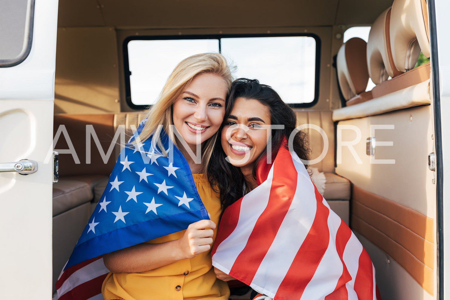 Two young female friends with American flag sitting in a camper van. Beautiful women with USA flag looking at camera.