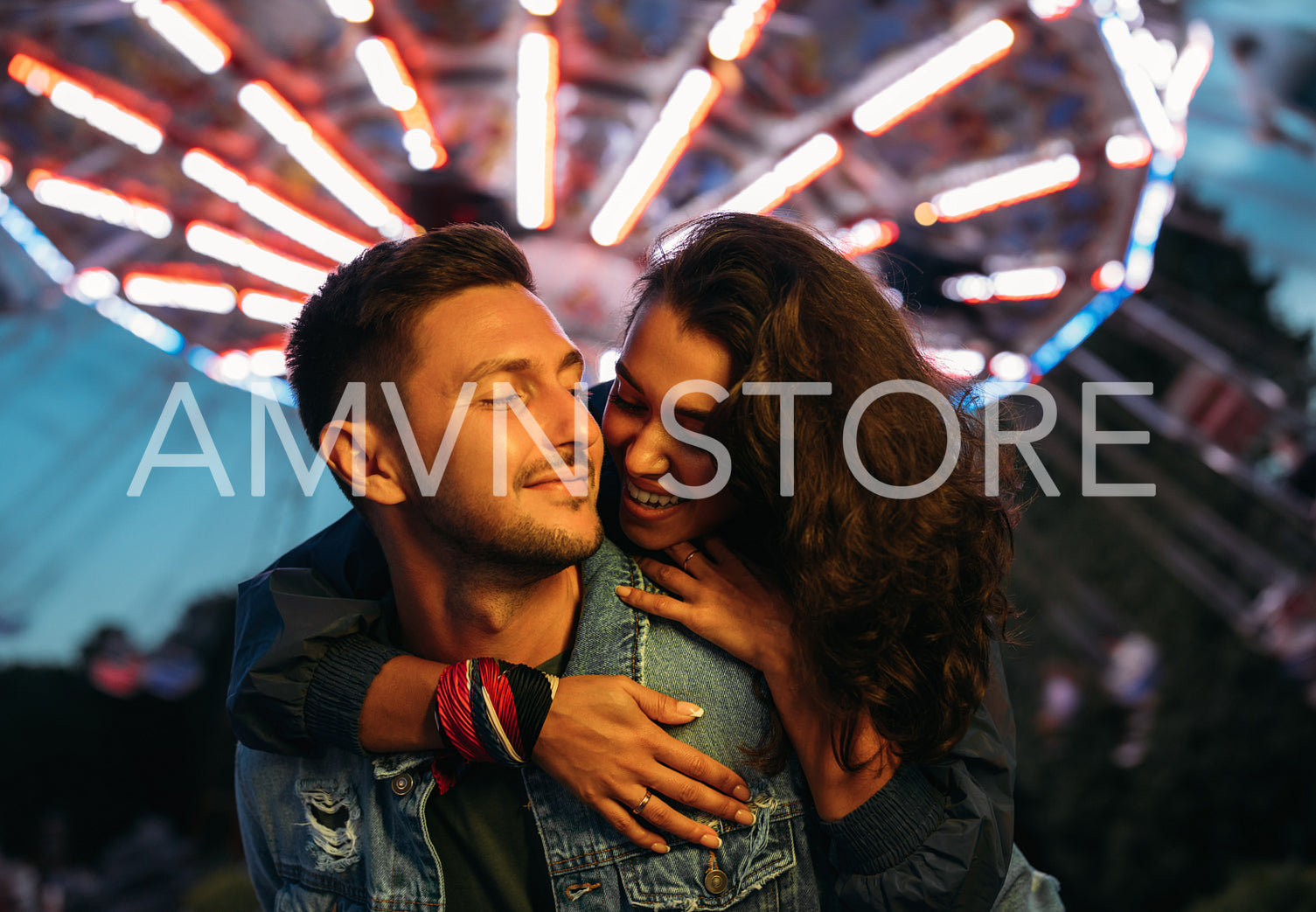 Smiling man piggybacks his happy girlfriend at the festival. Young couple having fun at night in an amusement park.