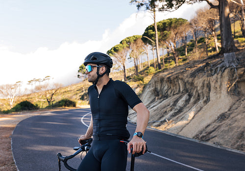 Side view of a professional biker relaxing leaning on his road bike. Male cyclist in glasses and helmet enjoying the view and taking a break during the ride.