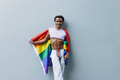 Happy guy with rainbow flag standing outdoors at grey wall and looking at camera wearing stylish clothes