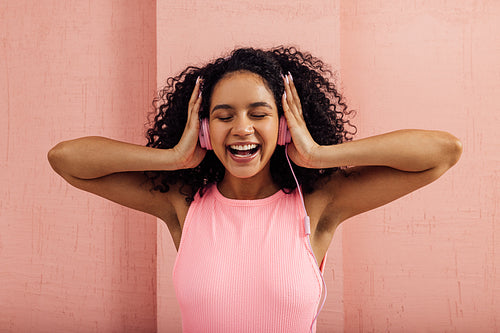 Laughing woman wearing pink headphones enjoying music with closed eyes. Smiling female with curly hair against a pink wall listening to music.