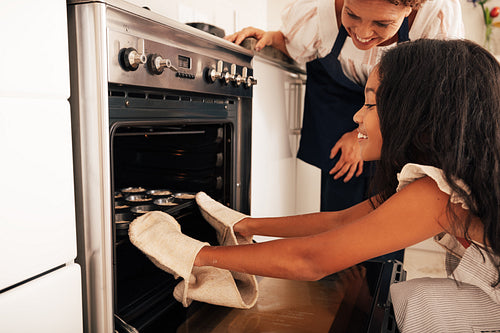 Granny and granddaughter putting unbaked cookies in oven. Girl wearing oven gloves putting cupcake molds.