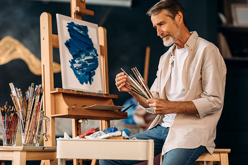 Mature painter holding a bunch of paint brushes sitting in studio