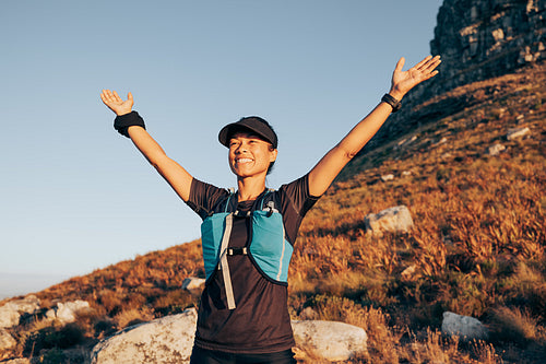 Happy woman enjoying sunset during hike. Smiling female standing on hill with raised arms.