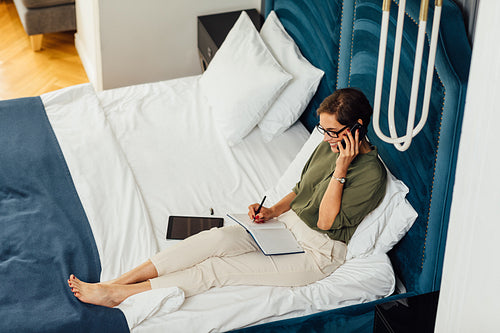 High angle view of woman in formal clothes lying on bed talking over mobile phone with a notebook on her hips